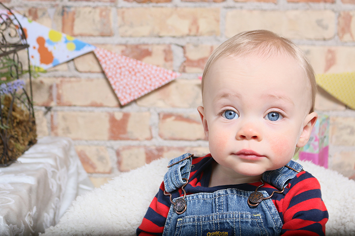 young boy wearing jean bib overalls and a blue and red striped shirt sitting against a brick wall